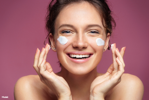 Joyful woman applying skincare cream to her cheeks and smiling