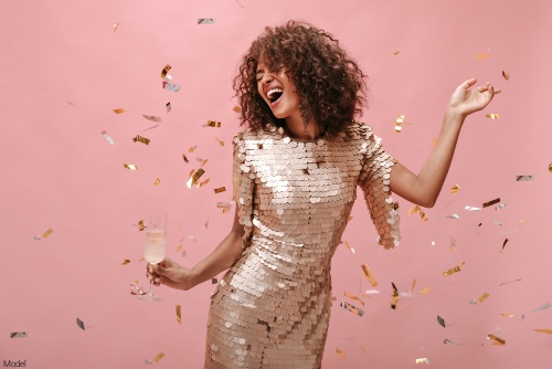 Joyful woman in a shimmery party dress holding a glass of champagne, dancing and smiling in front of a pink background as confetti falls around her