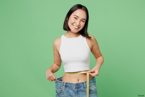 Slim woman smiling while measuring her waist with a fabric tape measure and showing off jeans that are too big