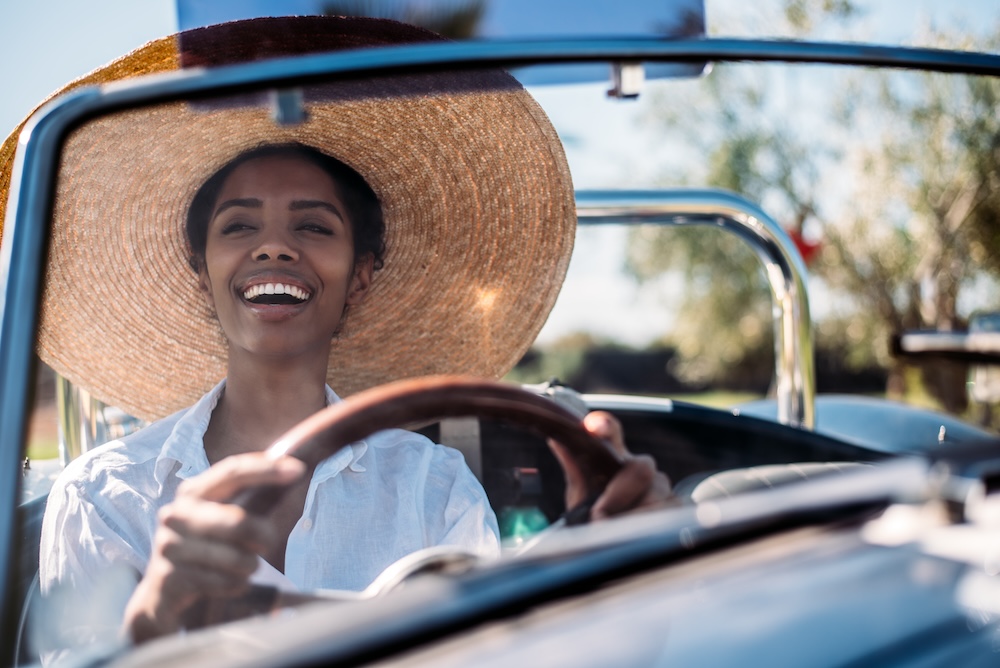 A woman of color wearing a large straw sunhat, smiling, driving a jeep with the top down.