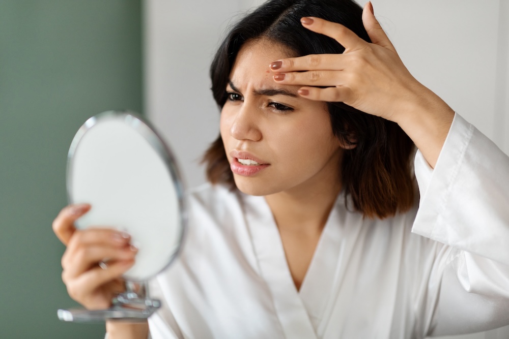 A woman with dark, cropped hair, scrunching her face while looking into a mirror, analyzing her acne scaring.