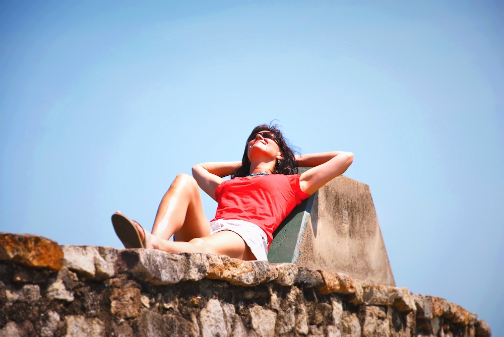 A woman in a red tee and khaki skirt lounging against a rock, sun-bathing in sunglasses.